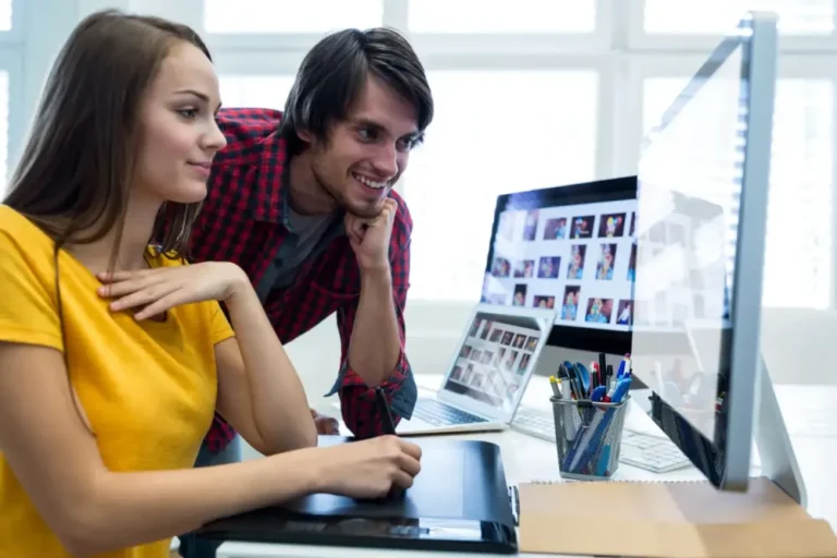 Two people collaborating on a web design project, with one person sitting at a desk using a graphic tablet and the other person standing and looking at the computer screen. The desk has a large monitor, a laptop displaying multiple images, and a container with pens and pencils.