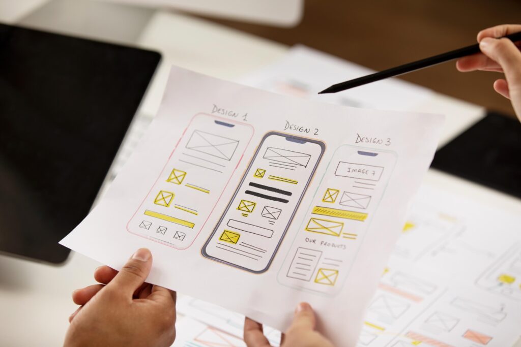 The image shows a person holding a sheet of paper with three different mobile app design mockups labeled "Design 1," "Design 2," and "Design 3." Each design features a variety of layout elements, such as placeholders for images, text blocks, and icons. Another person is pointing at the designs with a pencil, indicating a discussion or evaluation of the design options. In the background, a tablet and other design sketches are partially visible on a desk.