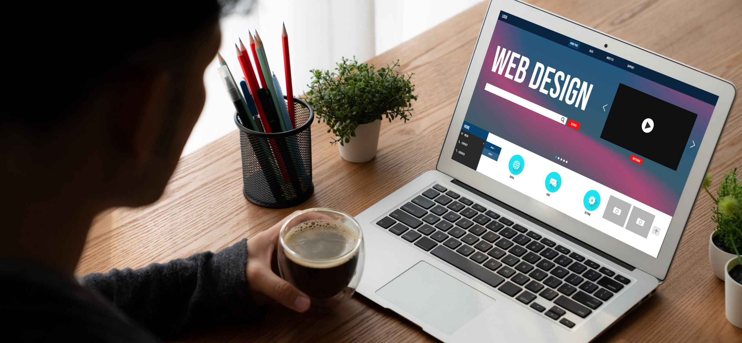 The image depicts an individual seated at a wooden desk, viewing a laptop screen displaying a webpage about web design. The webpage features icons for optimization, responsive design, and other web development tools. The person holds a cup of coffee, and there are pencils in a holder and a potted plant on the desk.